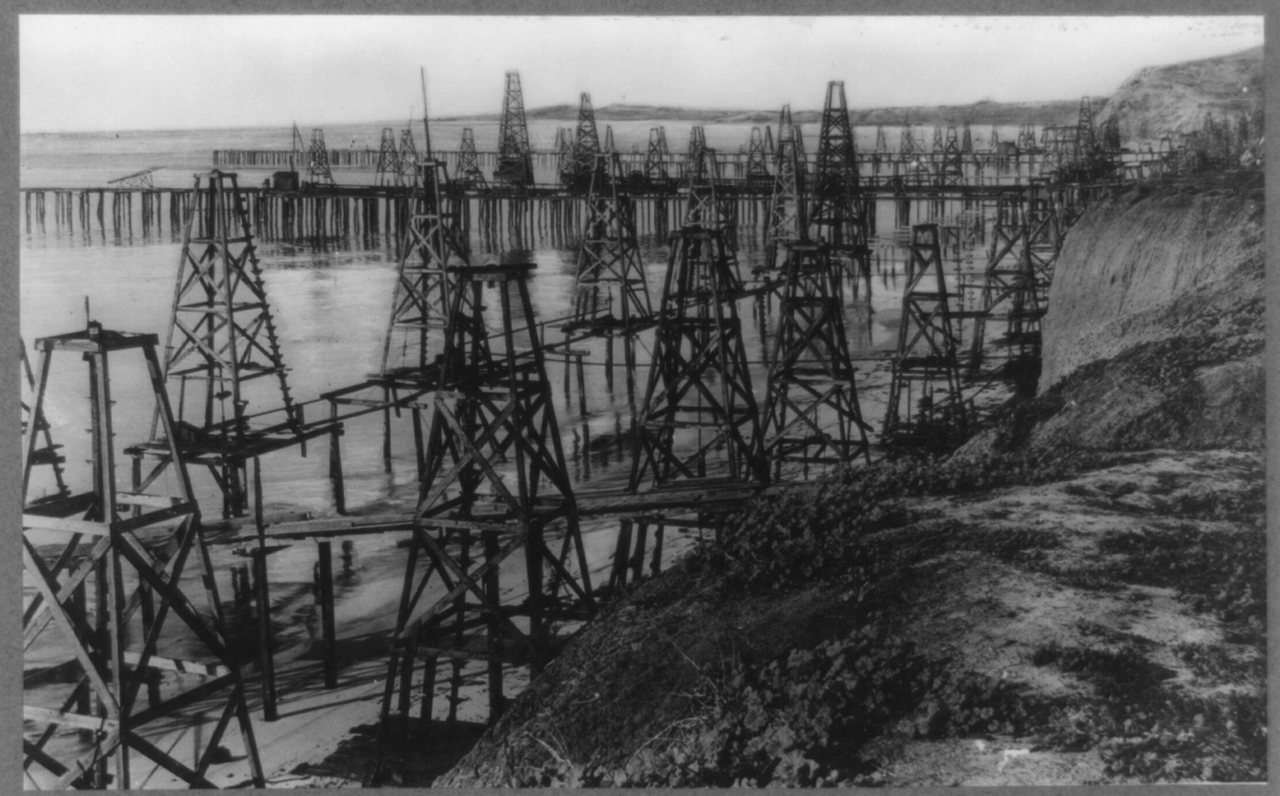 Dozens of wooden oil derricks standing on pile-supported piers over shallow water, extending from an eroding bluff out into the ocean at Summerland, California.
